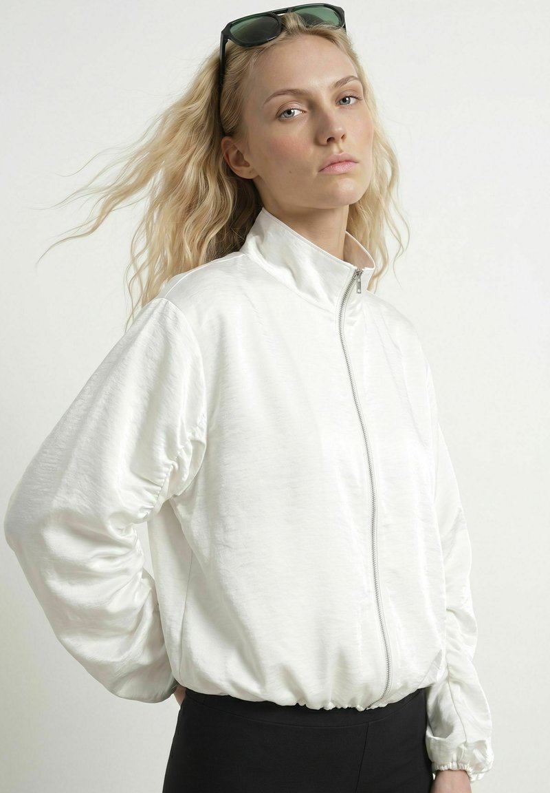 Femme blonde avec des lunettes de soleil sur la tête porte une veste blanche zippée et un pantalon noir, debout devant un fond clair uni.