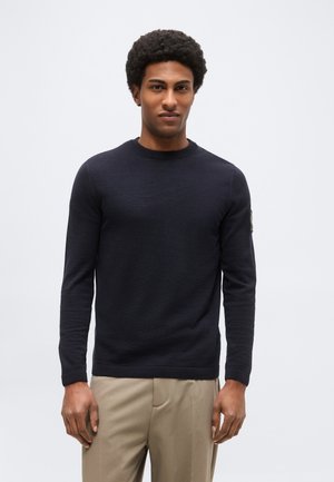 Muž v priliehavom čiernom pulóvri s dlhým rukávom a béžových nohaviciach stojí pred jednoliatym svetlosivým pozadím.