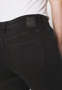 Detalle de una persona que lleva jeans de mezclilla negros con un bolsillo trasero y una etiqueta de la marca "PIECES" en la cinturilla.