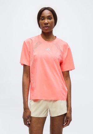 Donna in piedi con indosso una maglietta sportiva a maniche corte di un brillante colore corallo con dettagli traforati e pantaloncini beige su sfondo semplice.