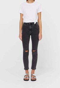 Černé skinny džíny s vysokým pasem a dírami na kolenou, doplněné o bílý uzlový tričko a stříbrné sandály. Hladká textura, přiléhavý design.