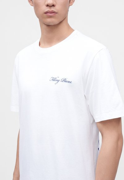 Άνδρας που φοράει απλό λευκό T-shirt με κοντά μανίκια και μικρό μπλε κείμενο με καλλιγραφικά γράμματα "Filling Pieces" στο στήθος.