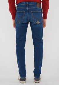 Uomo che indossa jeans blu con tasche posteriori e sneakers bianche, in piedi contro uno sfondo grigio uniforme, con le mani rilassate ai lati.