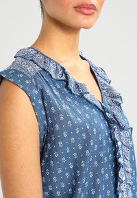 Modrá bezrukávová blůzka s volánkovým límcem, s bílou květinovou potiskem a složitými paisley detaily podél výstřihu. Měkká textilie.