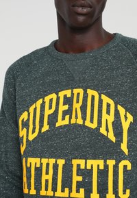Sudadera gris con tejido texturizado, con un destacado estampado amarillo "SUPERDRY ATHLETIC" en la parte frontal. Diseño clásico de cuello redondo.