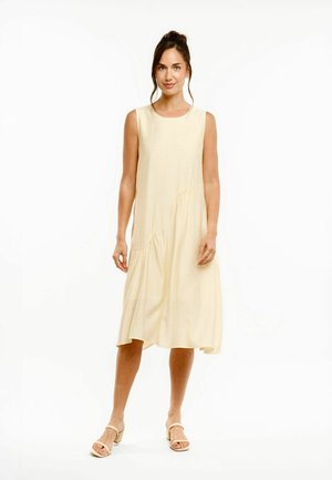Kvinde står iført en ærmeløs, lys beige midi-kjole med subtile samlinger og beige åbne blokhælssandaler.