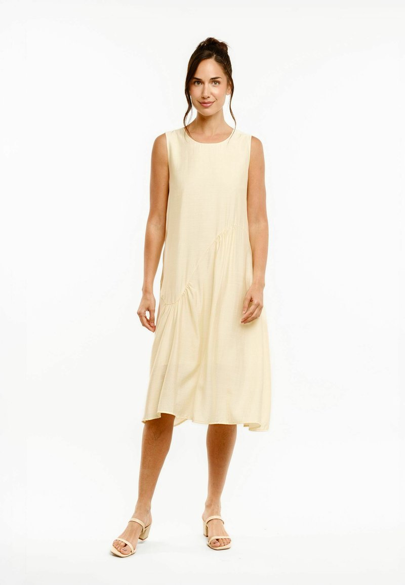 Femme debout portant une robe midi sans manches beige clair avec des détails subtilement froncés et des sandales beiges à talons blocs ouverts à l'avant.