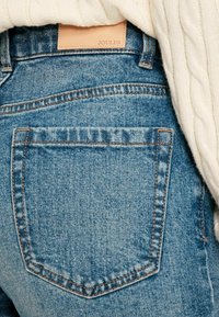 Plave traperice s klasičnim krojem, stražnjim džepom s detaljima šivanja i kožnom etiketom s natpisom "JOULES" na pojasu.