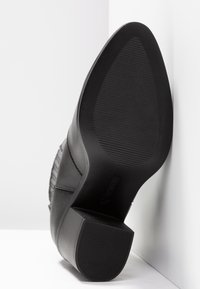 Chaussure en cuir noir avec une surface texturée, un bout rond et un talon de hauteur moyenne. La semelle présente un design côtelé pour une meilleure adhérence.
