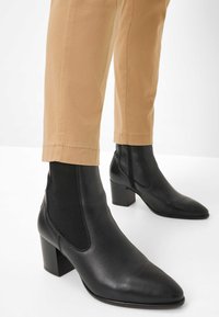 Bottines en cuir noir à bout pointu et à talon block, avec des panneaux élastiques sur les côtés et une texture lisse, accompagnées de pantalons beiges.
