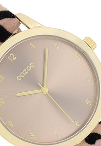 Montre dorée avec un cadran rond, un cadran dégradé beige, des index horaires dorés et un bracelet en tissu à motif avec des taches noires.