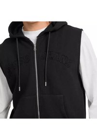 Sudadera sin mangas negra con cierre de cremallera, bolsillos frontales y bordado texturizado de "Guess Jeans" en el pecho. Tejido suave con un corte relajado.