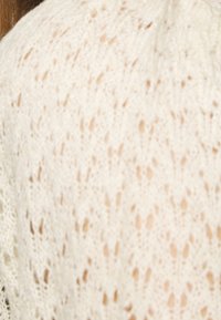 Biały, teksturowany dzianinowy materiał z otwartym wzorem koronki, ukazujący drobne szczegóły ściegów oraz miękki, lekki wygląd.