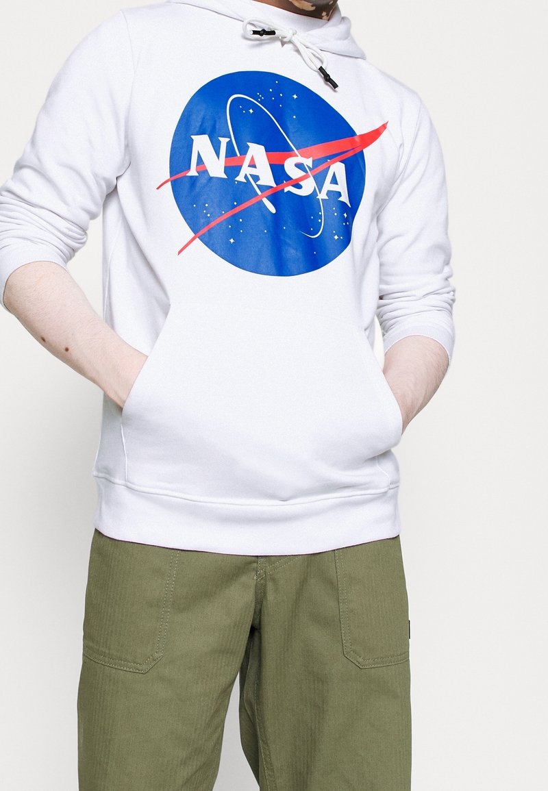 Sweat à capuche blanc arborant un logo NASA bleu avec un design accentué de rouge. La capuche est équipée de cordons de serrage et d'une poche avant. Porté avec un pantalon vert olive.