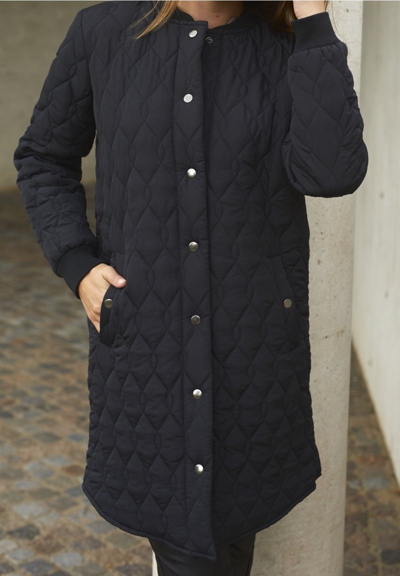 Cappotto lungo nero trapuntato con scollo rotondo e bottoni a pressione, dotato di tasche laterali e polsini elasticizzati, realizzato in tessuto morbido e texturizzato.