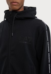 Sudadera con cremallera negra hecha de un tejido suave, que cuenta con capucha, bolsillo tipo canguro, logotipo tonal de EA7 en el pecho y cinta de marca a lo largo de las mangas.