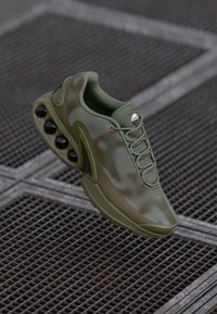 Olive green athletic shoe with textured upper, prominent laces, and a distinctive sole featuring five black air pockets. Reflective logo accent.