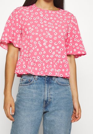 Vrouw draagt een roze bloemige korte mouwen blouse met ruchtrand, gecombineerd met een hoog getailleerde blauwe spijkerbroek.