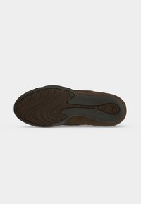 Semelle d’une chaussure marron avec une surface en caoutchouc noir et gris texturée antidérapante et un logo circulaire près du talon sur un fond uni.
