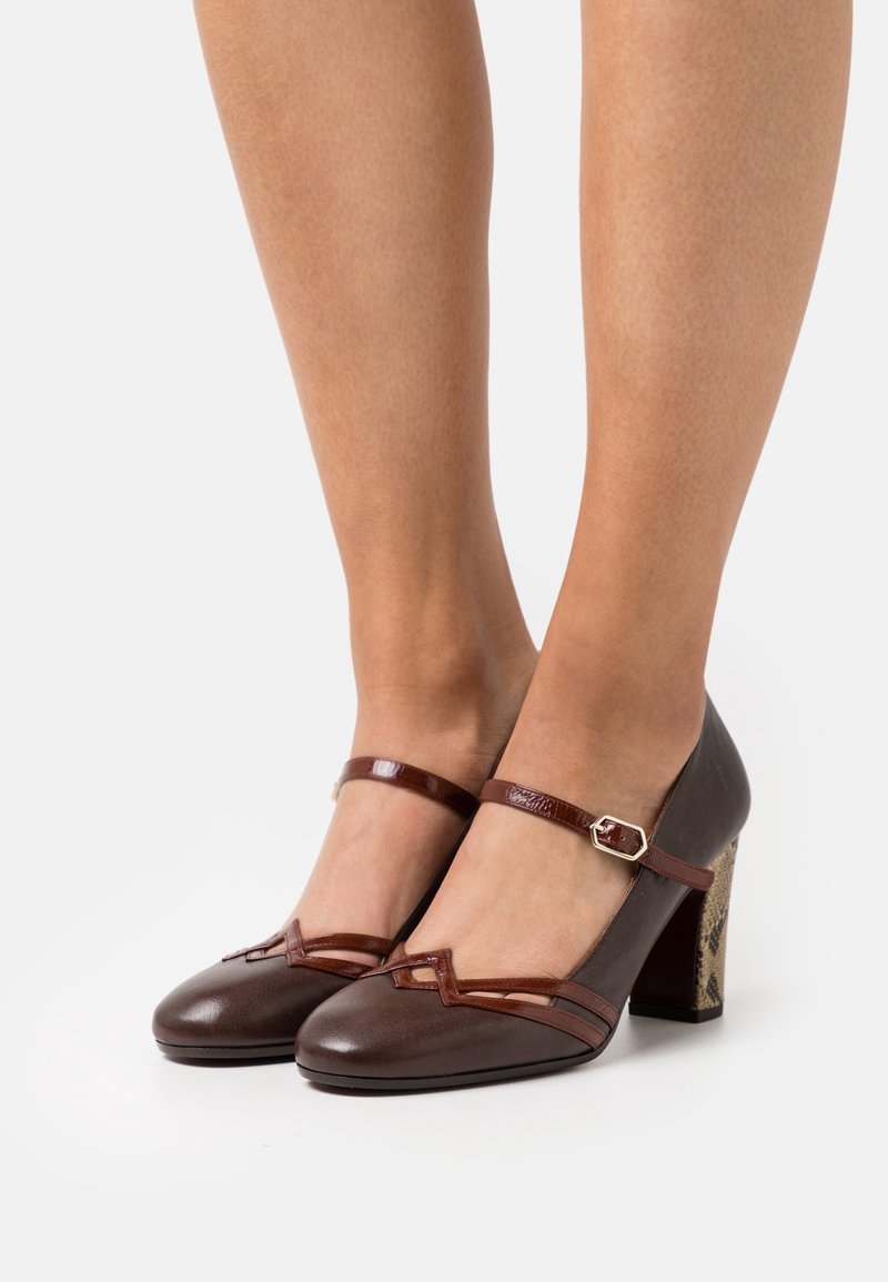 Talons hauts en cuir marron avec un bout carré, une sangle de cheville et un talon bloc à motif. Présente des accents brillants marron contrastants.