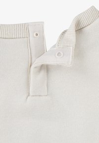 Tessuto a maglia beige con colletto ribattuto, dotato di due bottoni a pressione e dettagli a coste. La texture è liscia con una leggera elasticità.