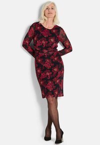 Schwarzes Kleid mit rotem Blumenmuster, langen transparenten Ärmeln, taillierter Silhouette und knielangem Saum. Kombiniert mit schwarzen High Heels.