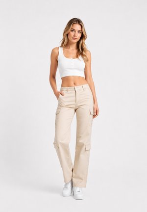 Junge Frau steht, trägt ein weißes bauchfreies Tanktop, beige Cargohosen und weiße Sneakers vor einem schlichten hellen Hintergrund.