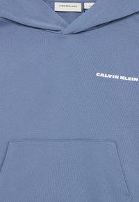 Hele sinine kapuutsiga särk, mis on valmistatud puuvillasegust, ja sellel on ees küljerihma tasku ning valge "Calvin Klein" logo. Sile tekstuur.