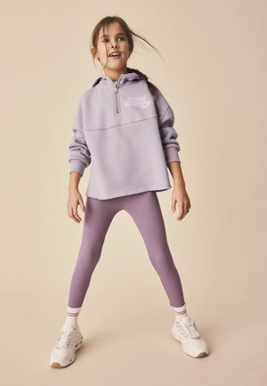 Young girl stands with legs apart wearing a lavender hoodie, matching leggings, white socks, and white sneakers against a beige background.