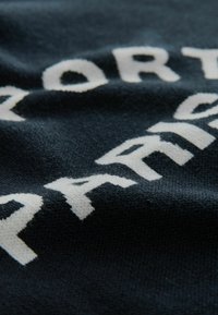 Úplet v tmavomodrej farbe s vyvýšeným bielym textom, na ktorom sú slová "SPORT" a "PARIS". Hladký povrch s jemným leskom.