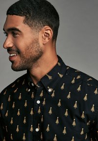 Chemise noire à boutons, ornée d'un motif d'arbre de Noël doré, avec un col étalé et des boutons blancs, présentée sur un modèle de côté.