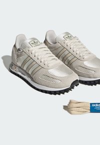 Ανοιχτό μπεζ αθλητικά παπούτσια Adidas με υφή σουέτ και ύφασμα, μαύρες σόλες με καρφιά, τρεις ρίγες και επιπλέον ταιριαστά κορδόνια δίπλα τους.