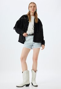 Zwarte suede shirt met franjes, zilveren studs, witte t-shirt, lichte denim shorts en witte cowboylaarzen met reliëfpatronen.