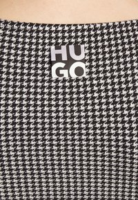 Schwarz-weißer Hahnentrittmuster-Stoff mit sichtbarem gewebtem Textur und einem auffälligen "HUGO" Markenlabel in kontrastierenden Farben.