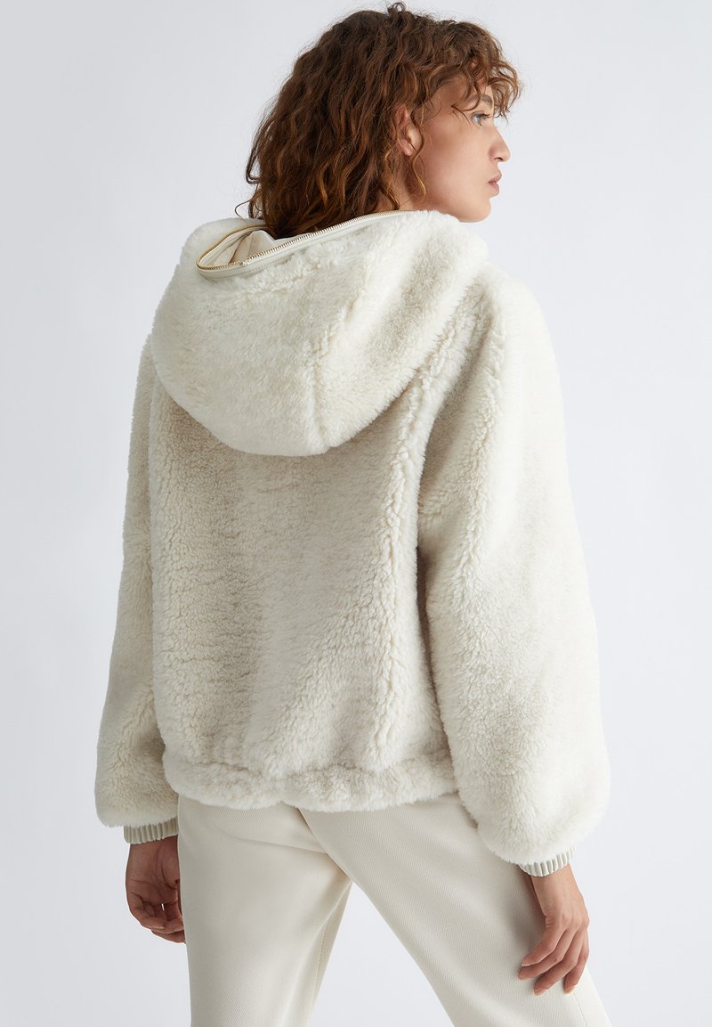 Hoodie en fausse fourrure crème avec une grande capuche, des épaules tombantes et des poignets côtelés. Texture douce, coupe décontractée, détail de fermeture éclair stylé au niveau du col.