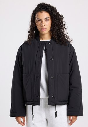 Veste matelassée noire avec un col rond, fermeture à boutons sur le devant, deux grandes poches, ourlet ajustable avec cordon de serrage et tissu texturé.