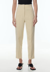 Hogewaist, beige broek van een gladde stof met zijzakken en een recht gesneden ontwerp; gedragen met zwarte haksandalen.