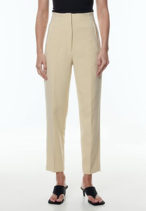 High-waisted, beige trousers made of smooth fabric with side pockets and a straight leg design; worn with black heeled sandals.