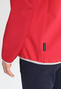 Rød jakke med glat, vandtæt stof, kontrasterende grå kant ved sømmen og manchetter, med et sort logomærke på sidesømmen.