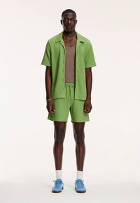 Rohelise tekstuuriga mustriga lühikeste varrukatega nööpidega särk, millele on sobitatud sobivad lühikesed püksid. All on pruun tank top ja jalas sinised spordijalatsid.