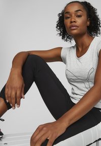 Tricou din bumbac gri cu logo-ul Puma negru, asociat cu colanți sportivi negri, ajustați. Modelul are părul creț și stă relaxat, cu brațele sprijinite.