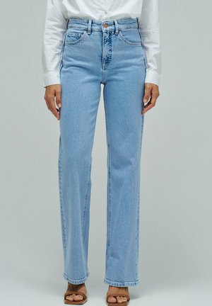 Personne portant un jean droit taille haute bleu clair, une chemise blanche boutonnée et des sandales beiges à bouts ouverts, debout devant un fond uni.