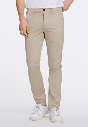 Mann trägt beige slim-fit Hose mit schwarzem Knopf, weiße Sneakers und ein in die Hose gestecktes weißes Hemd, steht mit einer Hand in der Tasche.