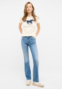 Camiseta crema con un gráfico de un caballo azul marino, combinada con jeans claros de campana. El modelo lleva zapatillas de sport beige. Diseño sencillo.