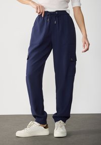Pantaloni cargo blu navy realizzati in tessuto liscio con tasche laterali e polsini elasticizzati. Presentano una vita con cordoncino per una vestibilità regolabile.