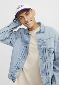 Lysblå denimjakke med knaplukning og sidelommer, båret over en beige skjorte. Beige kasket med blå skrift vist.
