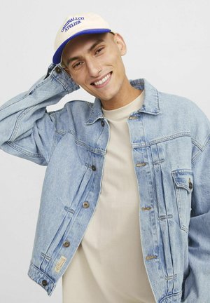 Lichtblauwe spijkerjack met knoopsluiting en zijzakken, gedragen over een beige shirt. Beige pet met blauwe tekst erop.