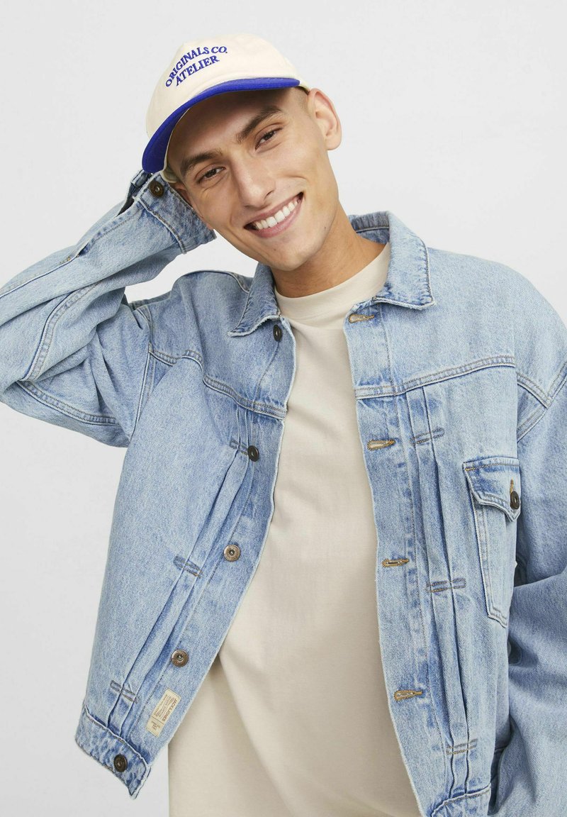 Lysblå denimjakke med knaplukning og sidelommer, båret over en beige skjorte. Beige kasket med blå skrift vist.