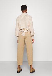 Maglione beige a maglia con maniche a palloncino, caratterizzato da un dettaglio annodato in vita, abbinato a pantaloni beige larghi e scarpe color miele.