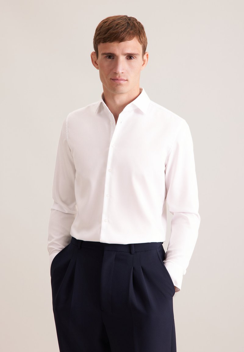 Homme aux cheveux bruns courts portant une chemise blanche boutonnée et un pantalon foncé, debout les mains dans les poches devant un fond uni.